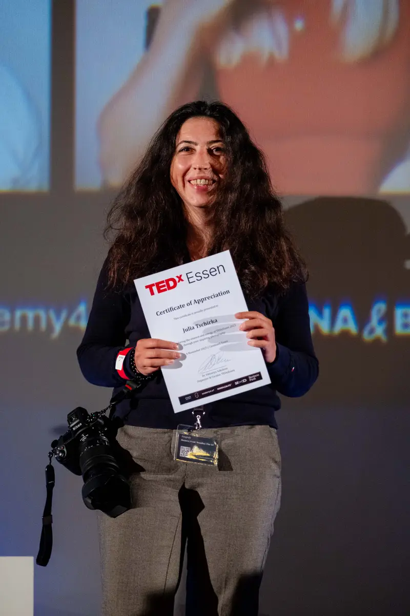 A guest smiles on stage holding her TEDxEssen certificate